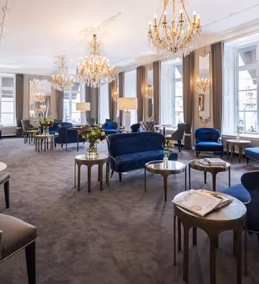 Elegant lounge med plysblå fløjlsstole, runde marmorborde og udsmykkede lysekroner. Store vinduer med flydende grå gardiner langs væggene, fylder rummet med naturligt lys. En luksuriøs og sofistikeret stemning hersker.
