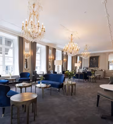 Elegant værelse med udsmykkede lysekroner, høje vinduer og klassisk indretning. Har plysblå fløjlsstole, små runde borde og dekorative spejle på lyse vægge. Et portrætmaleri hænger i baggrunden og forstærker de luksuriøse rammer.