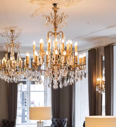 Elegant værelse med flere udsmykkede lysekroner, der hænger fra loftet og reflekterer lyset hen over rummet. Rummet har grå gardiner, hvide vægge og et stort spejl. Der kommer blødt naturligt lys ind fra vinduerne.