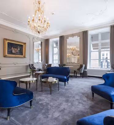En luksuriøs lounge med elegante blå fløjlsstole, en lysekrone og store vinduer med grå gardiner. Et guldindrammet maleri på væggen komplementerer den raffinerede indretning, og friske blomster tilføjer et strejf af farve til de sofistikerede omgivelser.