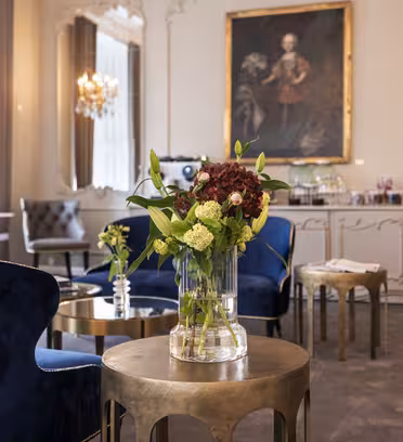 Elegant værelse med blå fløjlsstole, borde med guld accent og en vase med røde og hvide blomster. Et stort portræt hænger på væggen. Krystallysekroner og store vinduer bidrager til rummets sofistikerede.