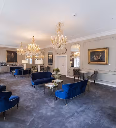Luksuriøs lounge med blå fløjlsstole, guldlysekroner og udsmykkede spejle. Rummet har plysgrå gulvtæpper og klassiske malerier på væggene. Små borde med blomsterarrangementer er spredt ud over, hvilket skaber en elegant atmosfære.