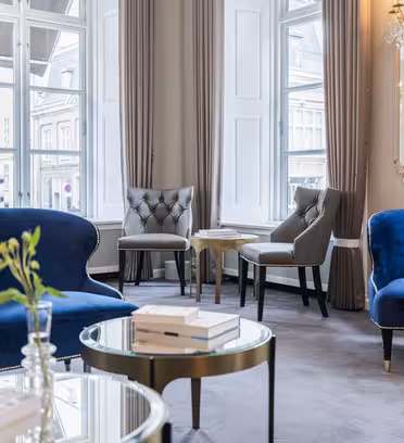 Elegant hotelobby med blå fløjlssofaer, grå polstrede stole og runde glassofaborde. Store vinduer med beige gardiner slipper rigeligt lys ind. Dekorative spejle og en lysekrone tilføjer sofistikering. Bøger og en plante pryder bordene.