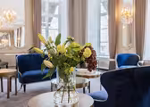 En luksuriøs stue har elegante blå fløjlsstole og en glasvase med frodige blomster på et rundt bord. Rummet er udsmykket med lysekroner og store vinduer, hvilket skaber en lys og sofistikeret atmosfære.