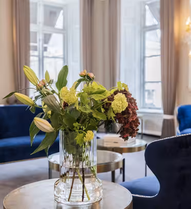 En luksuriøs stue har elegante blå fløjlsstole og en glasvase med frodige blomster på et rundt bord. Rummet er udsmykket med lysekroner og store vinduer, hvilket skaber en lys og sofistikeret atmosfære.