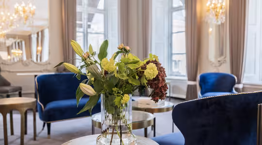 En luksuriøs stue har elegante blå fløjlsstole og en glasvase med frodige blomster på et rundt bord. Rummet er udsmykket med lysekroner og store vinduer, hvilket skaber en lys og sofistikeret atmosfære.