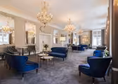 Elegant lounge med luksuriøse blå fløjlsstole og sofaer, udsmykkede lysekroner og store dekorative spejle på væggene. Rummet har et klassisk design med gulvtæppe og blødt naturligt lys fra høje vinduer.