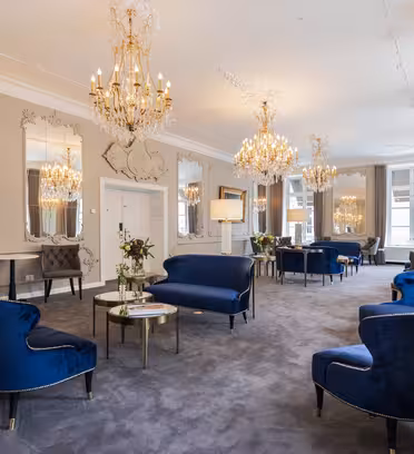 Elegant lounge med luksuriøse blå fløjlsstole og sofaer, udsmykkede lysekroner og store dekorative spejle på væggene. Rummet har et klassisk design med gulvtæppe og blødt naturligt lys fra høje vinduer.