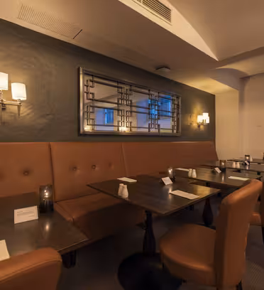 Et hyggeligt, svagt oplyst restaurantinteriør med brune lædersæder og mørke træborde. Små stearinlys og borddækning tilføjer stemning. Et stort dekorativt spejl og væglamper forstærker den varme atmosfære.
