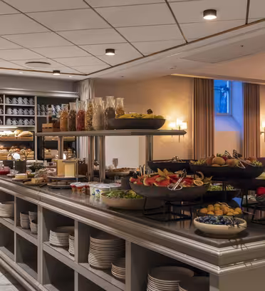 En buffet på en restaurant byder på skåle med frisk frugt og grøntsager, krukker med korn og forskellige retter. Det veloplyste interiør har et moderne design med hylder, tallerkener og en hyggelig atmosfære.