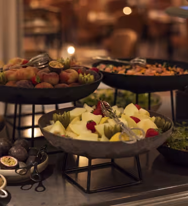 Et buffetbord med forskellige retter, herunder kiwi, figner, æbler, ferskner og blandet grønt. Frugterne er arrangeret i sorte skåle, og rammerne har en varm, indbydende stemning med blød belysning.