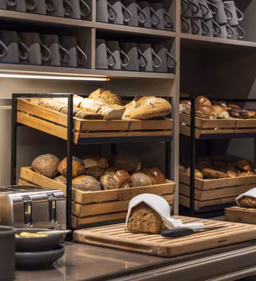 En bagerudstilling med træbakker fyldt med forskellige brød, herunder brød og rundstykker. Et skærebræt med et snittet brød og kniv er i forgrunden. Hylderne ovenover rummer kaffekopper. Belysningen er varm og hyggelig.