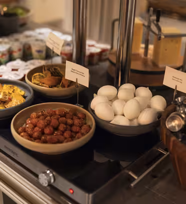 En buffetopstilling med skåle indeholdende retter som kogte æg, frikadeller, skiveskåret ost og chips. Der er også små kopper yoghurt og en beholder med redskaber i nærheden.