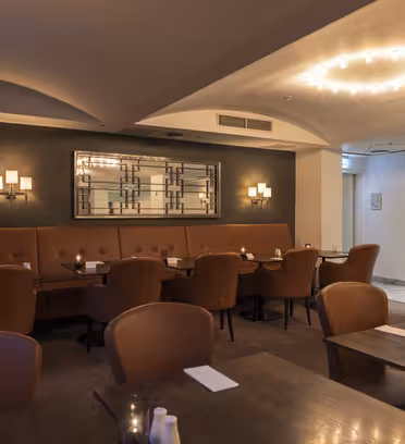 Moderne restaurantinteriør med brune læderstole og borde, svagt oplyst af væglamper og cirkulære loftslamper. Et stort dekorativt spejl hænger på en mørk accentvæg, hvilket skaber en hyggelig og intim atmosfære.