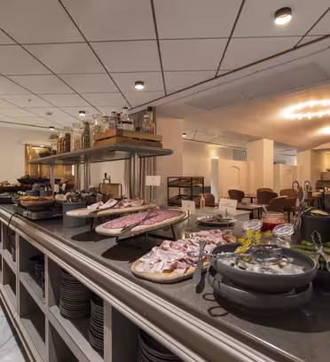 En buffet med forskellige kød, oste, grøntsager og krydderier er sat op på en bordplade i et veloplyst spiseområde. Borde og stole er arrangeret i det rummelige, elegant designede værelse med blød overheadbelysning.