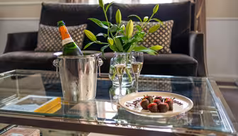 Et sofabord af glas med en flaske champagne i en isspand, to fyldte glas, en tallerken med chokoladeovertrukne jordbær og en vase med liljer, placeret i en hyggelig stue med en mørk sofa og mønstrede puder.