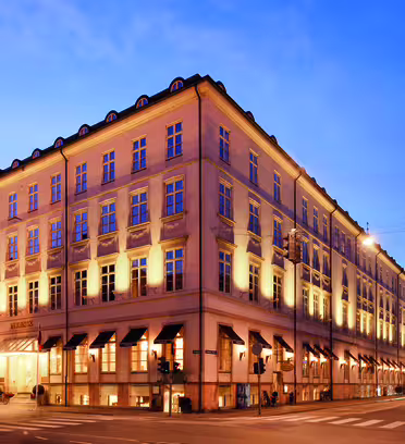 En stor, elegant bygning med varme lys, der oplyser facaden i skumringen. Det har en klassisk arkitektonisk stil med flere vinduer og en hjørneindgang. Himlen er klar, og gaden foran er tom.