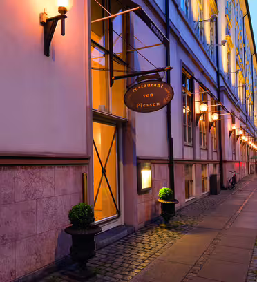 Gadebillede af en bygning med et restaurantskilt "restaurant von Plessen" oplyst af varme lys i skumringen. Stien er brolagt med brosten, og små potteplanter flankerer indgangen. En cykel hviler mod bygningen i baggrunden.