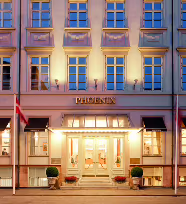 Billedet viser facaden på Hotel Phoenix med dets beige ydre og ensartede vinduer. "PHOENIX" vises tydeligt over indgangen, som er flankeret af potteplanter og to flag. Bygningen er varmt oplyst.
