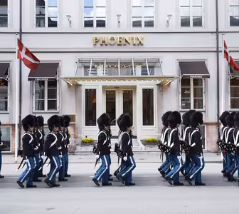 Et optog af uniformerede vagter i sorte hatte og blå bukser marcherer foran Phoenix-hotellet, med danske flag vist på begge sider af indgangen.