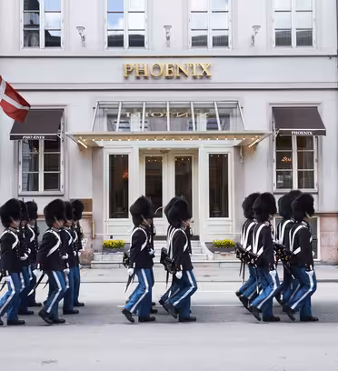Et optog af uniformerede vagter i sorte hatte og blå bukser marcherer foran Phoenix-hotellet, med danske flag vist på begge sider af indgangen.
