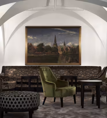 Elegant lounge med hvælvede hvide lofter, mønstrede stole og en central grøn lænestol. Et stort landskabsmaleri af en kirke med en flod og træer hænger på væggen. Sorte borde er placeret mellem siddepladserne.