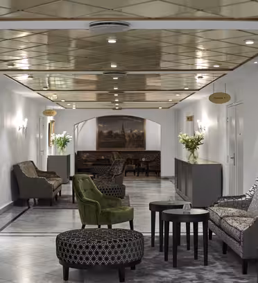 En hotellounge med marmorgulv og et reflekterende loft. Rummet byder på elegante grå og grønne møbler, indrammede malerier på væggene og vaser med hvide blomster. Der er arrangeret borde og lænestole på begge sider.