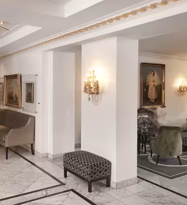 Luksuriøs hotellets lobby med elegant marmorgulv og stilfulde siddepladser. Vægge prydet med klassisk kunst og oplyst af udsmykkede væglampetter. En storslået trappe med et guldrækværk er i baggrunden.
