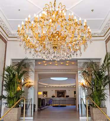 Luksuriøs hotelindgang med en storslået krystallysekrone, marmorvægge, spejlede accenter og to store potteplanter. Et opholdsområde og reception er synlige i baggrunden.