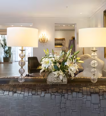 Elegant hotellobby med en sølvmønstret sofa, to krystalbaselamper med hvide skærme og en vase med hvide liljer. Kunstværker og udsmykkede væglamper pryder væggene og skaber en sofistikeret stemning.