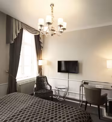 Et stilfuldt hotelværelse med en dobbeltseng med en mønstret dyne, et vindue med gardiner, et væghængt tv, en lille lysekrone, et glasbord med en stol og et skrivebord med en lampe og stol. Neutrale toner og moderne indretning fuldender rammerne.