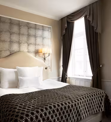 Et hyggeligt hotelværelse med en pænt lavet dobbeltseng med et sort/hvidt mønstret tæppe og hvide puder. To væglamper giver varm belysning over sengen. Elegante mørke gardiner indrammer et lyst vindue til højre.