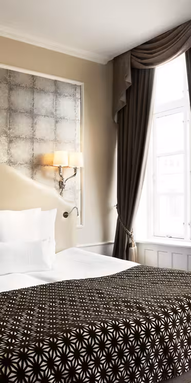 Et hyggeligt hotelværelse med en pænt lavet dobbeltseng med et sort/hvidt mønstret tæppe og hvide puder. To væglamper giver varm belysning over sengen. Elegante mørke gardiner indrammer et lyst vindue til højre.