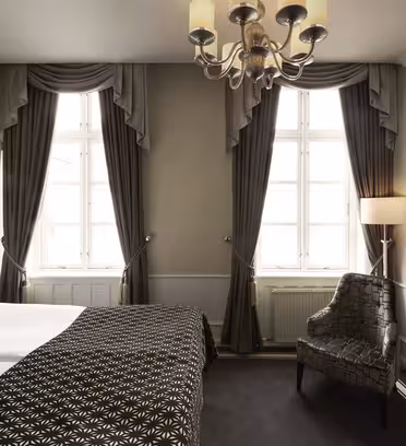 Elegant hotelværelse med dobbeltseng, mønstret mørkebrunt tæppe og hvide puder. To høje vinduer med kraftige gardiner lukker sollys ind. En polstret stol og et sidebord i glas sidder tæt på et vægmonteret tv. En lysekrone hænger fra loftet.