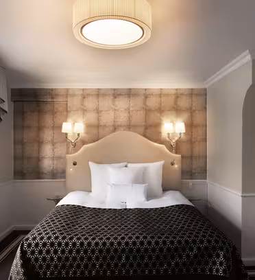 Et hyggeligt hotelværelse med en stor seng dækket af et mønstret mørkebrunt og hvidt tæppe. Rummet har elegante væglamper, et rundt loftslys, et lille skrivebord ved et vindue med draperede gardiner og en vægmonteret knagerække.