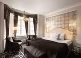 Luksuriøst hotelværelse med en king-size seng med en polstret sengegavl, flankeret af glaslamper. Et opholdsområde ved store vinduer omfatter to lænestole og en fodskammel. Draperer og en lysekrone tilføjer elegance til rummet. Neutral farvepalet hele vejen igennem.