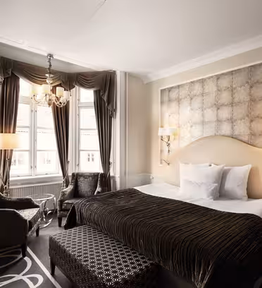 Luksuriøst hotelværelse med en king-size seng med en polstret sengegavl, flankeret af glaslamper. Et opholdsområde ved store vinduer omfatter to lænestole og en fodskammel. Draperer og en lysekrone tilføjer elegance til rummet. Neutral farvepalet hele vejen igennem.
