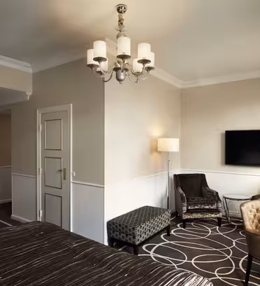Elegant hotelværelse med stor seng, mønstret tæppe og væghængt tv. Rummet har en lysekrone, lænestole med glasbord, en mønstret bænk og to gulvlamper. Væggene er neutrale, og vinduet er beklædt med gardiner.
