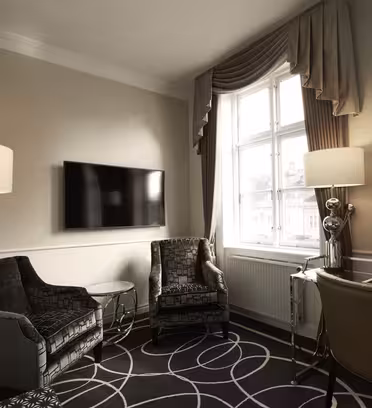 Et stilfuldt hotelværelse har to mørke lænestole med mønstret stof, et glasbord, et skrivebord med en stol, en gulvlampe og et væghængt tv. Stort vindue med gardiner slipper naturligt lys ind og oplyser det mønstrede tæppe.