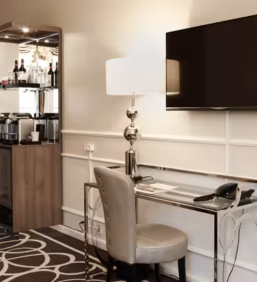 Hotelværelseshjørne med skrivebord, stol og telefon i glas under et væghængt tv. En minibar med kaffemaskine og drikkevarer er i baggrunden ved siden af en stående lampe med hvid skærm. Tæppet har et cirkulært mønster.