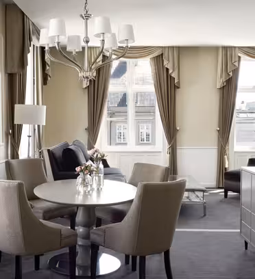 En lys, elegant indrettet stue med beige vægge og gardiner. Den har et rundt bord med fire stole, en sort sofa, en lysekrone og store vinduer, der giver rigeligt naturligt lys. En kommode og dekorative elementer fremhæver den raffinerede stil.