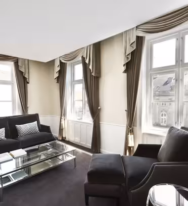 Sofistikeret stue med to store vinduespartier indrammet af elegante brune gardiner. Værelset har en sort sofa, to lænestole, et glas sofabord og en elegant gulvlampe, der skaber en moderne og luftig atmosfære.