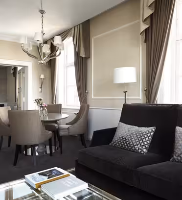 Elegant hotelsuite med opholdsstue med sort sofa og glassofabord, der støder op til en spiseplads med et rundt bord og fire stole. Et soveværelse med en stor seng er synlig gennem en åben døråbning. Neutrale farver og blød belysning fremhæver indretningen.