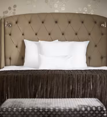 Et luksuriøst hotelværelse med en plys, tuftet beige sengegavl med hvide puder og sengetøj. Sengen har et brunt, fløjlsblødt sengetæppe. To moderne væglamper og sideborde med blomstret tapetbagtæppe fuldender det elegante look.
