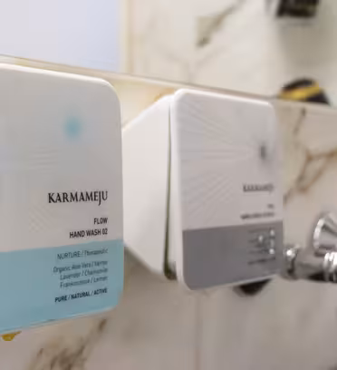 Nærbillede af to vægmonterede sæbedispensere i et badeværelse med marmorbagtæppe. Den venstre er mærket "Karmameju Flow Hand Wash 02" med et lyseblåt design, og den højre har et lignende design med en anden etiket.