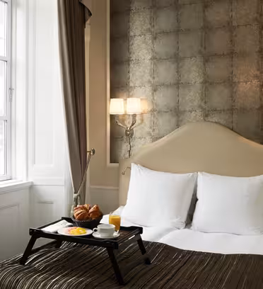 Et hyggeligt hotelværelse med en dobbeltseng, hvide puder og en metallisk tekstureret sengegavl. En morgenmadsbakke med kager, juice og frugt er på sengen. To væglamper oplyser rummet. Et vindue med gardiner giver naturligt lys.