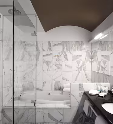 Et moderne badeværelse med marmor flisebelagt væg og gulv. Den har en glasindkapslet bruser, et badekar, en sort bordplade med toiletartikler og håndklæder pænt arrangeret. Belysningen er blød, hvilket skaber en ren og elegant atmosfære.