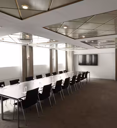 Et moderne mødelokale med et langt rektangulært bord omgivet af sorte stole. Værelset har et spejlloft og beige gardiner, med et stort fladskærms-tv monteret på en hvid væg for enden.