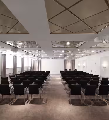 Et moderne, tomt mødelokale med rækker af sorte stole mod en hvid skærm foran. Rummet har et geometrisk loftdesign med firkantede paneler, beige tæppe og lyse beige vægge med lampetter. Store vinduer med gardiner langs den ene side.