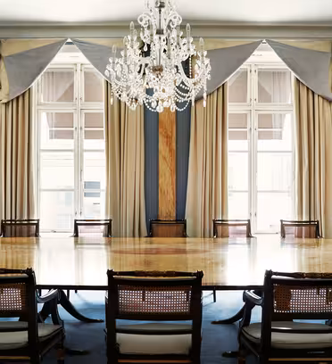 Udsmykket mødelokale med et poleret træbord, omgivet af elegante stole. Store vinduer er prydet med luksuriøse blå og beige gardiner, og en storslået lysekrone hænger fra loftet og tilføjer et strejf af sofistikering.